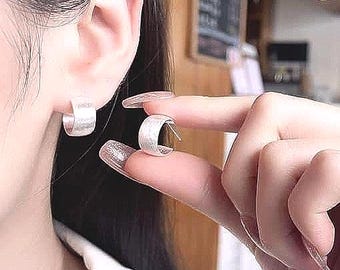 May include: A pair of silver hoop earrings with a textured surface. One earring is being held up to an ear, while the other is already in place. The earrings have a simple, modern design.
