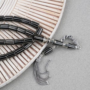 Peut inclure: Un chapelet composé de perles cylindriques noires avec de fines rayures verticales blanches. Les perles sont reliées par des éléments en métal argenté, incluant une pièce centrale décorative et un pompon en chaîne argentée à plusieurs brins.