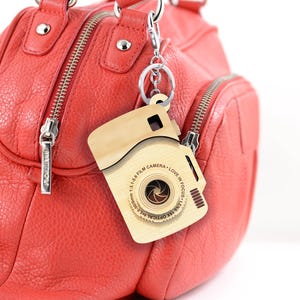 May include: A wooden camera keychain with the text "5.0-90/0mm 1:3.1-5.6 FILM CAMERA - LOVE IN FOCUS - LENS 184 OPTICAL 15.0" engraved on it. The keychain is attached to a red leather handbag.
