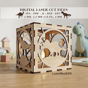 May include: A wooden laser-cut dinosaur-themed lantern. The cube-shaped lantern features dinosaur silhouettes and circular cutouts. The text "DIGITAL LASER CUT FILES" and "LaserBeeSVG" are visible. The lantern is 3.2 mm (1/8 in) thick.