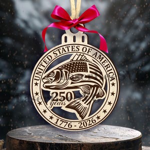 Może przedstawiać: Drewniana ozdoba choinkowa z motywem ryby, amerykańską flagą i napisem "UNITED STATES OF AMERICA 250 years 1776-2026". Okrągła ozdoba ma czerwoną wstążkę i beżowy sznurek do zawieszenia.
