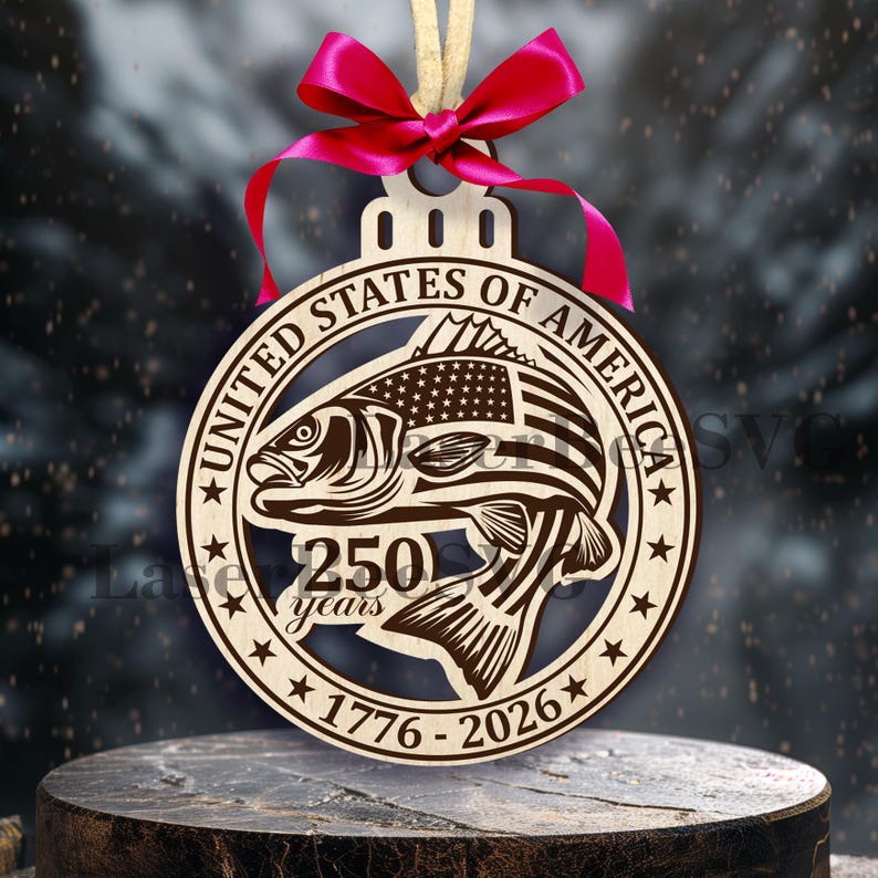 Może przedstawiać: Drewniana ozdoba choinkowa z motywem ryby, amerykańską flagą i napisem "UNITED STATES OF AMERICA 250 years 1776-2026". Okrągła ozdoba ma czerwoną wstążkę i beżowy sznurek do zawieszenia.
