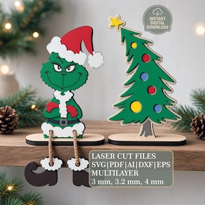 May include: A green character wearing a red and white Santa hat and suit with dangling legs, alongside a green Christmas tree with a yellow star and colorful ornaments. Text reads: LASER CUT FILES SVG|PDF|AI|DXF|EPS MULTILAYER 3 mm, 3.2 mm, 4 mm.