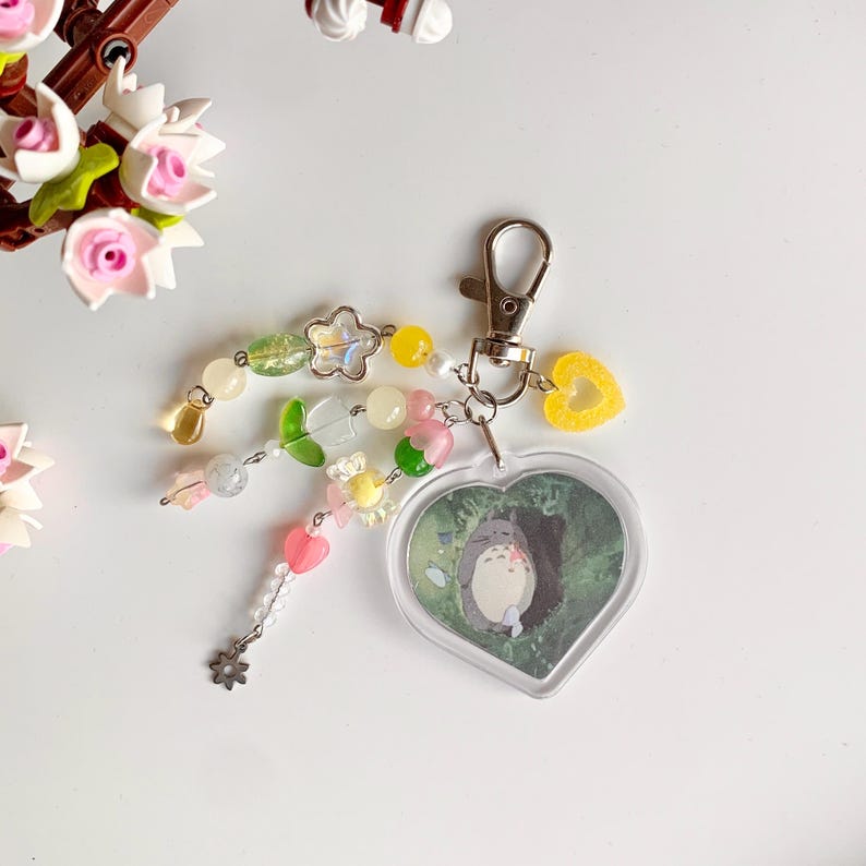 May include: A heart-shaped keychain with a clear plastic charm featuring a cartoon character from the movie My Neighbor Totoro. The charm is surrounded by a colorful beaded chain with green, yellow, pink, and white beads.