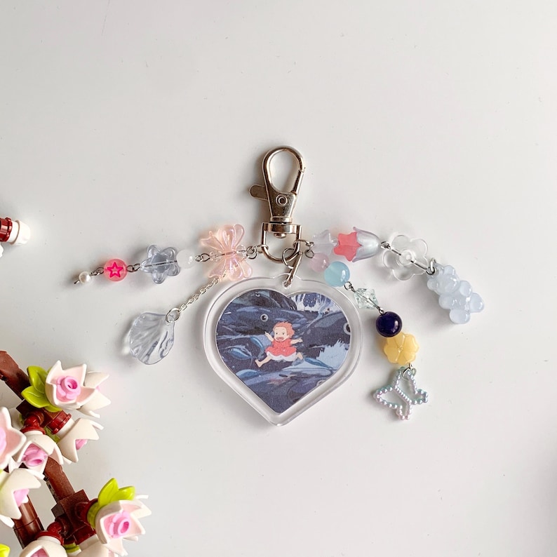 May include: A heart-shaped keychain with a picture of a girl in a red dress standing on a rock in a river. The keychain is decorated with beads, charms, and butterflies in various colors, including pink, blue, and yellow.