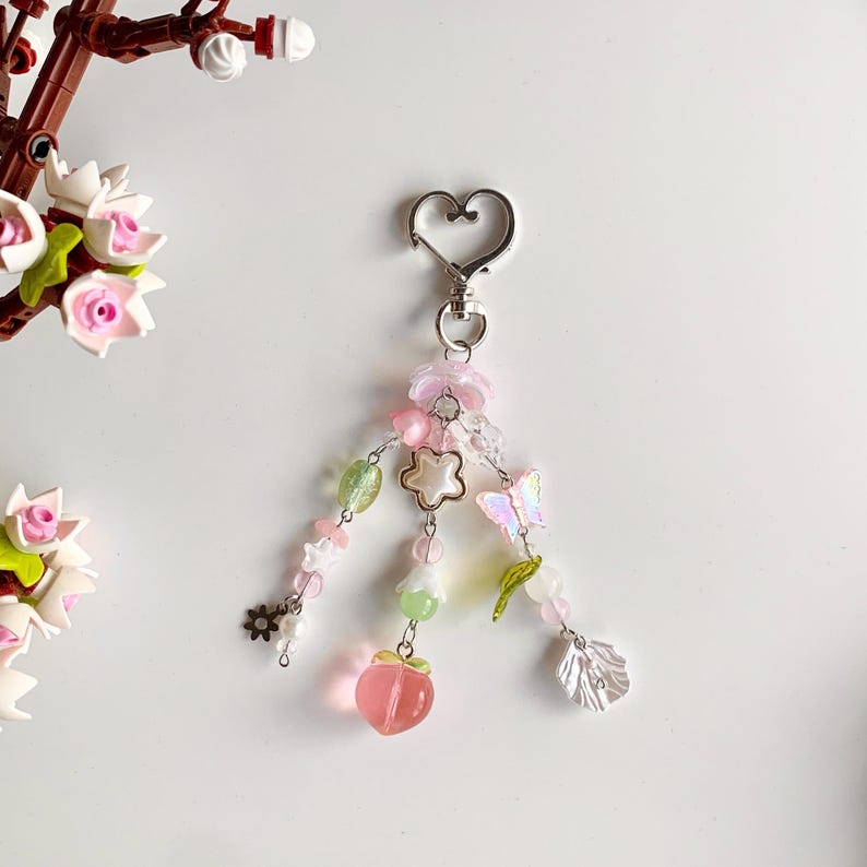 May include: A pink and green charm with a peach, butterfly, and flower accents. The charm is attached to a silver heart-shaped keychain.
