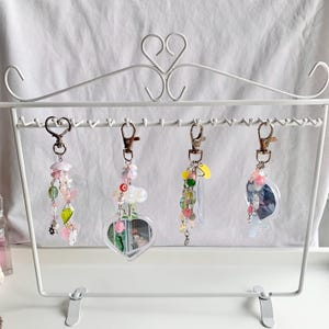 May include: Four colorful charm keychains with different designs hanging on a white metal display stand. Each keychain features a variety of beads and charms, including hearts, stars, and flowers.