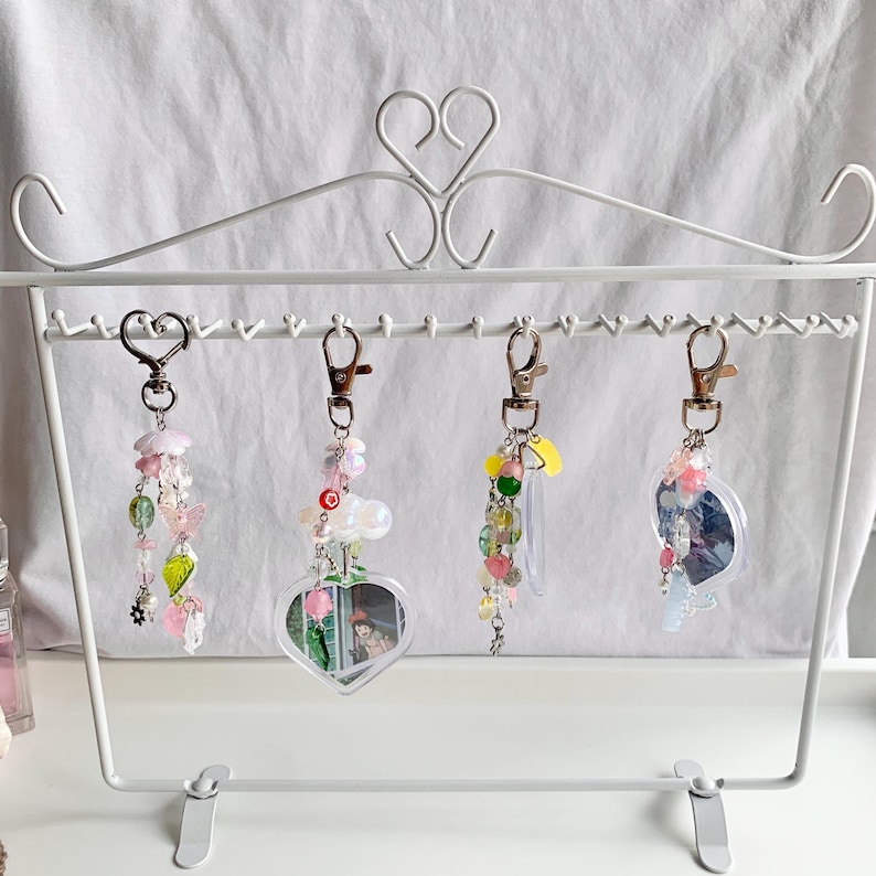 May include: Four colorful charm keychains with different designs hanging on a white metal display stand. Each keychain features a variety of beads and charms, including hearts, stars, and flowers.