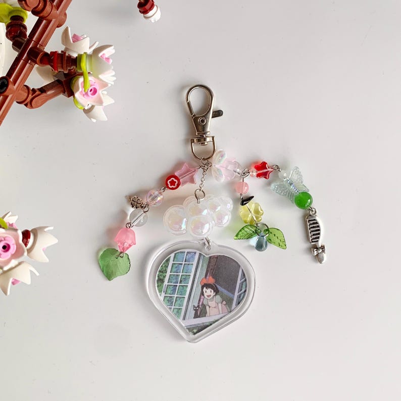 May include: A heart-shaped charm with a picture of a girl looking out a window, attached to a silver chain with colorful beads, charms, and a key.