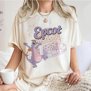 May include: A cream colored t-shirt with a purple cartoon dragon and the text "Epcot" in purple. The dragon is standing in front of a purple and white outline of the Spaceship Earth attraction at Epcot.