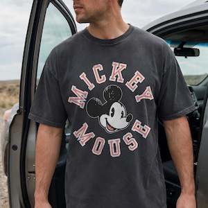May include: A charcoal gray t-shirt featuring a distressed Mickey Mouse graphic. The design includes the words "Mickey Mouse" in a circular arrangement with red accents. The shirt is paired with dark blue jeans.