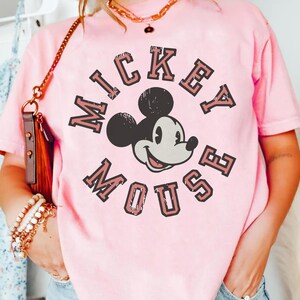 May include: A light pink t-shirt featuring a graphic of Mickey Mouse. The text "Mickey Mouse" is arched around the character's face in a vintage, collegiate style. The shirt has a crew neck and short sleeves.