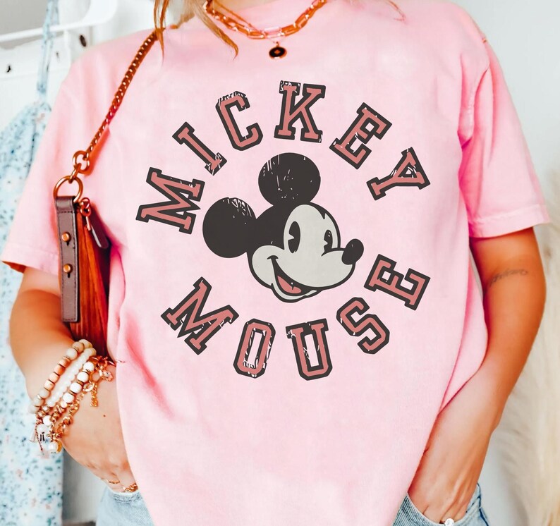 May include: A light pink t-shirt featuring a graphic of Mickey Mouse. The text "Mickey Mouse" is arched around the character's face in a vintage, collegiate style. The shirt has a crew neck and short sleeves.