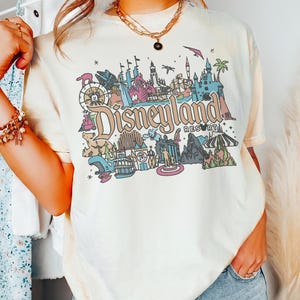 May include: Cream-colored t-shirt featuring a colorful Disneyland Resort graphic. The graphic includes various park icons, the Disneyland Resort logo, and a vintage aesthetic. The shirt is a relaxed fit.