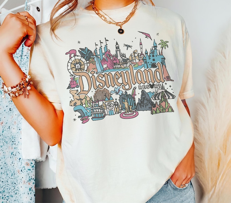May include: Cream-colored t-shirt featuring a colorful Disneyland Resort graphic. The graphic includes various park icons, the Disneyland Resort logo, and a vintage aesthetic. The shirt is a relaxed fit.