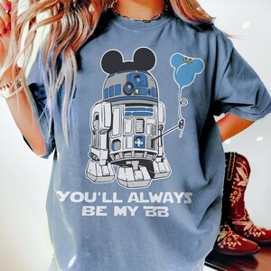 May include: A blue t-shirt featuring a graphic of R2-D2 with Mickey Mouse ears and a blue balloon. The text "YOU'LL ALWAYS BE MY BB" is printed below the graphic. The shirt is a light blue color.
