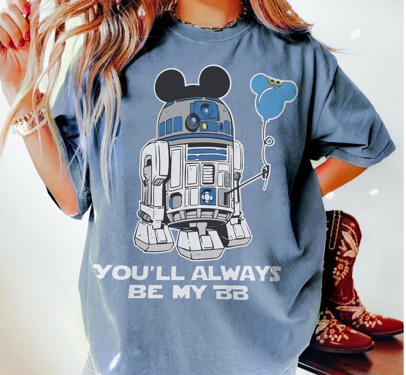 May include: A blue t-shirt featuring a graphic of R2-D2 with Mickey Mouse ears and a blue balloon. The text "YOU'LL ALWAYS BE MY BB" is printed below the graphic. The shirt is a light blue color.