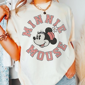 Puede incluir: Camiseta color crema con un gr&aacute;fico de Minnie Mouse y el texto "MINNIE MOUSE" en una fuente rosa retro. Minnie Mouse se representa con un lazo rosa y una flor blanca. La camiseta tiene mangas cortas y un corte holgado.