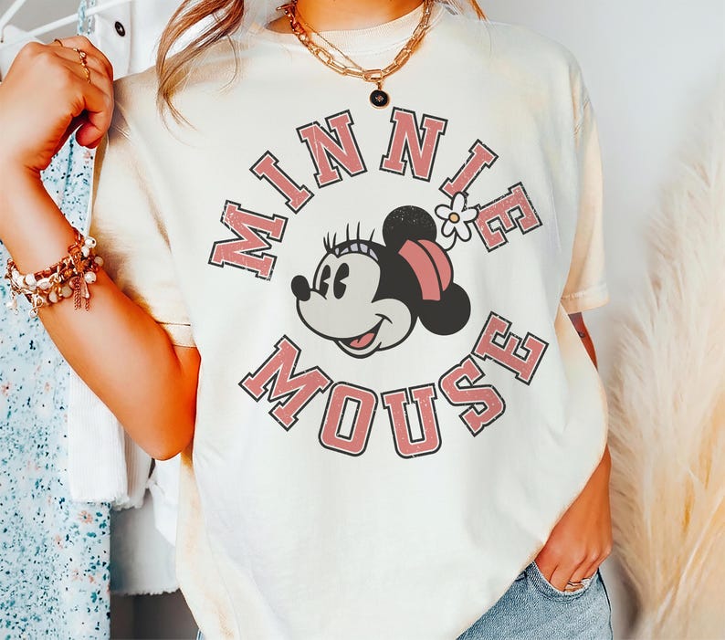 Puede incluir: Camiseta color crema con un gr&aacute;fico de Minnie Mouse y el texto "MINNIE MOUSE" en una fuente rosa retro. Minnie Mouse se representa con un lazo rosa y una flor blanca. La camiseta tiene mangas cortas y un corte holgado.