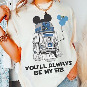 May include: Cream-colored t-shirt featuring a graphic of R2-D2 with Mickey Mouse ears and a blue balloon. The text "YOU'LL ALWAYS BE MY BB" is printed below the graphic. The shirt has short sleeves.