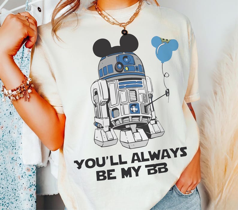 May include: Cream-colored t-shirt featuring a graphic of R2-D2 with Mickey Mouse ears and a blue balloon. The text "YOU'LL ALWAYS BE MY BB" is printed below the graphic. The shirt has short sleeves.