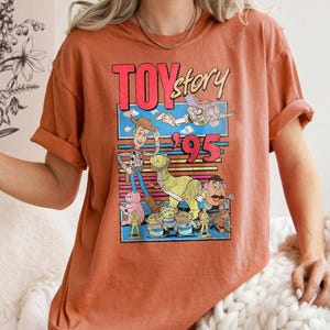 May include: Burnt orange t-shirt featuring a colorful graphic from the movie "Toy Story." The graphic includes the text "Toy Story '95" and various cartoon characters. The shirt has short sleeves and a relaxed fit.