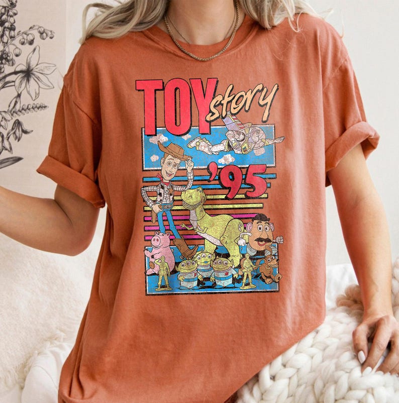 May include: Burnt orange t-shirt featuring a colorful graphic from the movie "Toy Story." The graphic includes the text "Toy Story '95" and various cartoon characters. The shirt has short sleeves and a relaxed fit.
