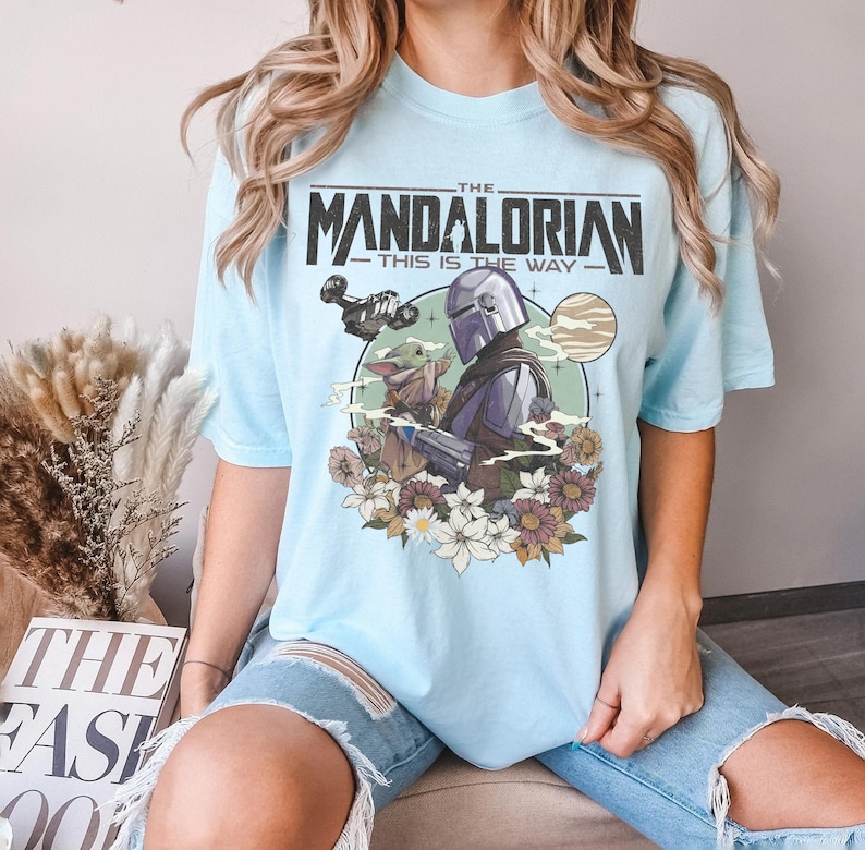 Puede incluir: Una camiseta de manga corta azul claro con un estampado gr&aacute;fico de The Mandalorian y Grogu. El dise&ntilde;o incluye el texto THE MANDALORIAN THIS IS THE WAY, una nave espacial, un planeta y un colorido arreglo floral en la base.