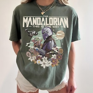 Puede incluir: Una camiseta verde oliva con un estampado gr&aacute;fico de The Mandalorian y Grogu. El texto dice THE MANDALORIAN THIS IS THE WAY. El dise&ntilde;o incluye una nave espacial, un planeta y un grupo de flores coloridas que rodean a los personajes.