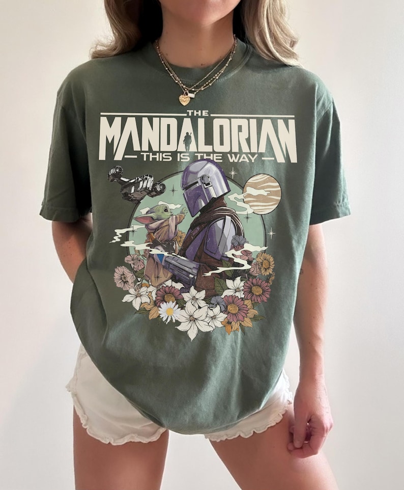 Puede incluir: Una camiseta verde oliva con un estampado gr&aacute;fico de The Mandalorian y Grogu. El texto dice THE MANDALORIAN THIS IS THE WAY. El dise&ntilde;o incluye una nave espacial, un planeta y un grupo de flores coloridas que rodean a los personajes.