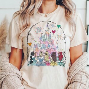 May include: A white t-shirt with a colorful illustration of Cinderella, Snow White, Ariel, Belle, and Rapunzel standing in front of a pink and blue watercolor castle with fireworks. The castle is surrounded by a black border with flowers and hearts.