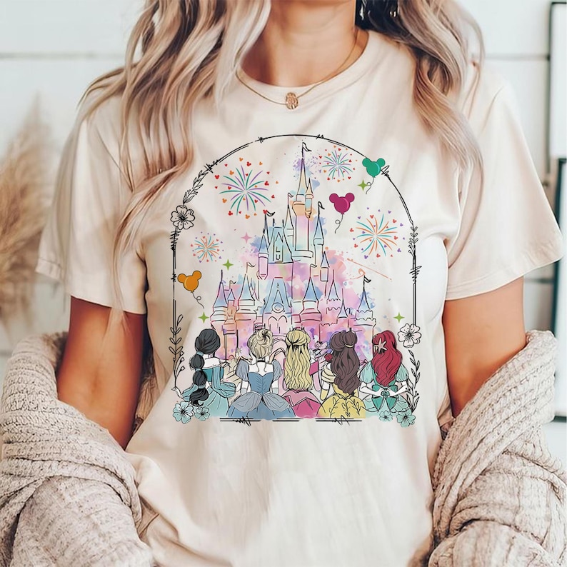 May include: A white t-shirt with a colorful illustration of Cinderella, Snow White, Ariel, Belle, and Rapunzel standing in front of a pink and blue watercolor castle with fireworks. The castle is surrounded by a black border with flowers and hearts.