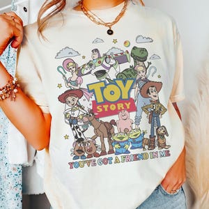 May include: Cream-colored t-shirt featuring a colorful graphic of Toy Story characters. The design includes Woody, Buzz Lightyear, and other characters from the movie, with the text "TOY STORY" and "YOU'VE GOT A FRIEND IN ME".
