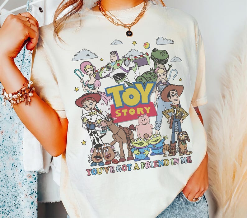 May include: Cream-colored t-shirt featuring a colorful graphic of Toy Story characters. The design includes Woody, Buzz Lightyear, and other characters from the movie, with the text "TOY STORY" and "YOU'VE GOT A FRIEND IN ME".