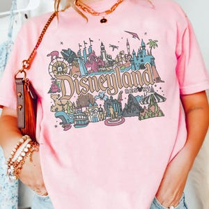 May include: Pink t-shirt with a colorful Disneyland Resort graphic. The design features various park icons, including a castle, a Ferris wheel, and the Disneyland Resort text. The shirt is a soft pink color.