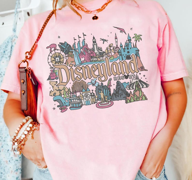 May include: Pink t-shirt with a colorful Disneyland Resort graphic. The design features various park icons, including a castle, a Ferris wheel, and the Disneyland Resort text. The shirt is a soft pink color.