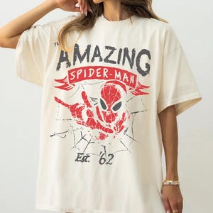 Pode incluir: Uma t-shirt de manga curta em tom off-white com uma estampa gr&aacute;fica do Spider-Man em vermelho e preto. O texto diz The Amazing Spider-Man com uma faixa vermelha e Est. '62 abaixo da personagem. O design inclui uma teia de aranha.