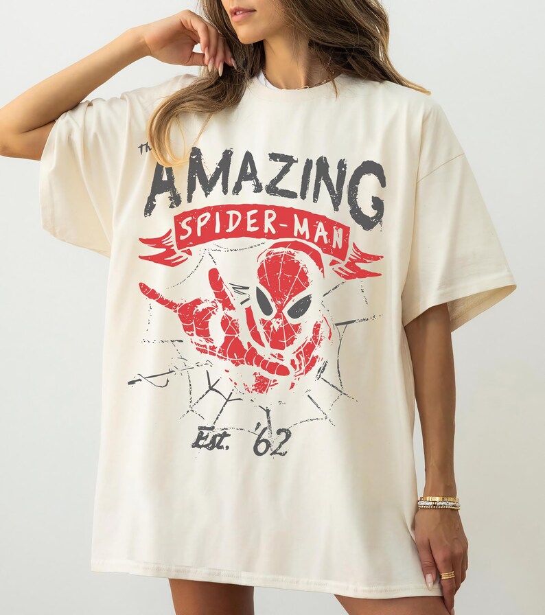 Pode incluir: Uma t-shirt de manga curta em tom off-white com uma estampa gr&aacute;fica do Spider-Man em vermelho e preto. O texto diz The Amazing Spider-Man com uma faixa vermelha e Est. '62 abaixo da personagem. O design inclui uma teia de aranha.