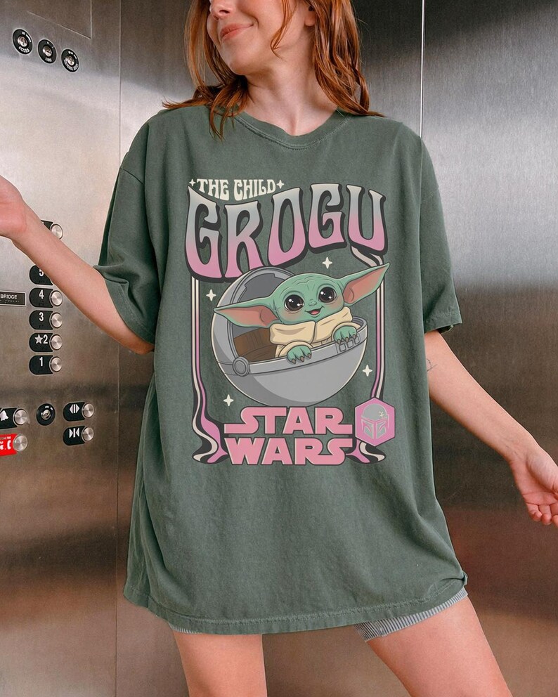 May include: A dark olive green short-sleeve t-shirt featuring a graphic print of the character Grogu from Star Wars inside a floating pod. The text reads The Child, Grogu, and Star Wars in a retro-style font with pink and white accents.