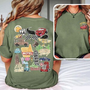 May include: Olive green t-shirt featuring a collection of cartoon car graphics and travel-themed stickers. The back of the shirt displays images of Route 66, checkered flags, and the words "Radiator Springs". The front of the shirt has a small graphic of a red car and the words "Piston Cup Champions".