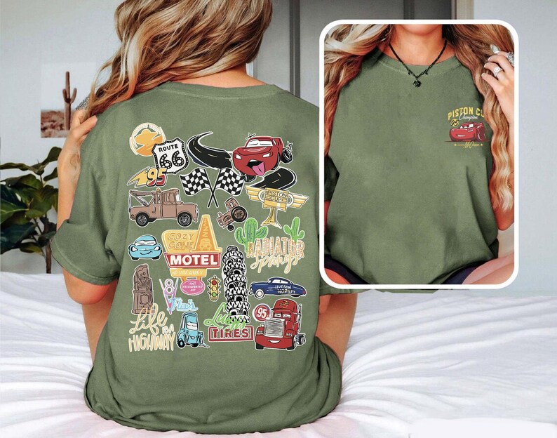 May include: Olive green t-shirt featuring a collection of cartoon car graphics and travel-themed stickers. The back of the shirt displays images of Route 66, checkered flags, and the words "Radiator Springs". The front of the shirt has a small graphic of a red car and the words "Piston Cup Champions".