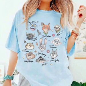 May include: A light blue t-shirt featuring a graphic print of various animated animal characters from Zootopia, including Judy Hopps, Nick Wilde, and Flash, with their names written in black script text alongside small star and icon accents.