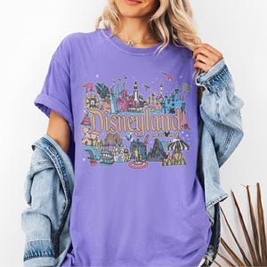 May include: A lilac-colored t-shirt featuring a colorful Disneyland Resort graphic. The graphic includes various park icons and the Disneyland Resort text. The shirt has short sleeves and a relaxed fit, with a denim jacket draped over the shoulders.