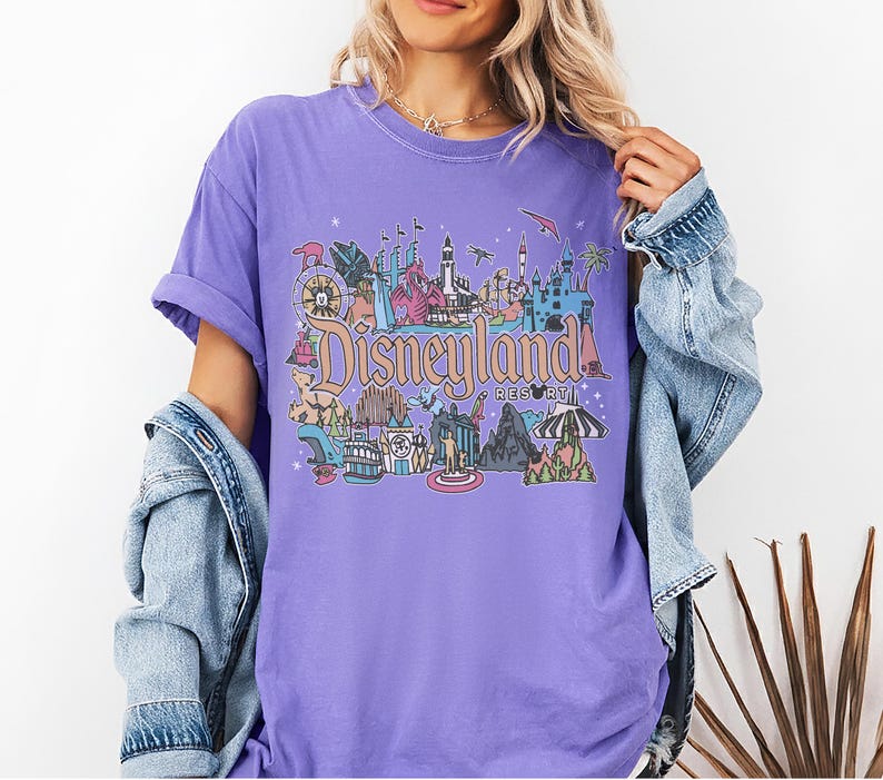 May include: A lilac-colored t-shirt featuring a colorful Disneyland Resort graphic. The graphic includes various park icons and the Disneyland Resort text. The shirt has short sleeves and a relaxed fit, with a denim jacket draped over the shoulders.