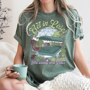 May include: Olive green t-shirt with the text "Get in Loser" in a whimsical font. The graphic features a boat ride surrounded by plants and the words "The Land" and "We're learning about Lettuce".