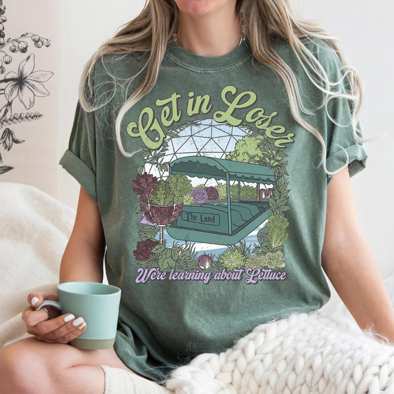 May include: Olive green t-shirt with the text "Get in Loser" in a whimsical font. The graphic features a boat ride surrounded by plants and the words "The Land" and "We're learning about Lettuce".