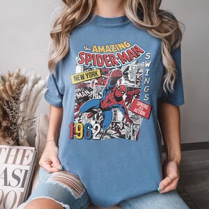 Peut inclure: Un t-shirt bleu avec un design de bande dessin&eacute;e vintage de Spider-Man. Le graphisme comprend le texte "The Amazing Spider-Man", "New York", "1962" et "Swings Spring Action". Le super-h&eacute;ros est repr&eacute;sent&eacute; dans son costume rouge et bleu classique.