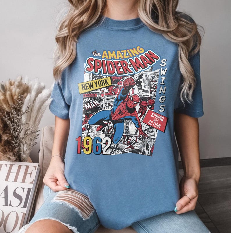 Peut inclure: Un t-shirt bleu avec un design de bande dessin&eacute;e vintage de Spider-Man. Le graphisme comprend le texte "The Amazing Spider-Man", "New York", "1962" et "Swings Spring Action". Le super-h&eacute;ros est repr&eacute;sent&eacute; dans son costume rouge et bleu classique.