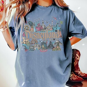 May include: A blue t-shirt featuring a colorful Disneyland Resort graphic. The graphic includes various landmarks and the Disneyland Resort logo. The shirt has short sleeves and a crew neck.