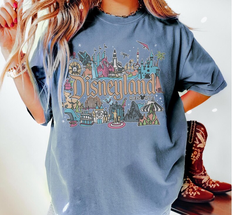 May include: A blue t-shirt featuring a colorful Disneyland Resort graphic. The graphic includes various landmarks and the Disneyland Resort logo. The shirt has short sleeves and a crew neck.